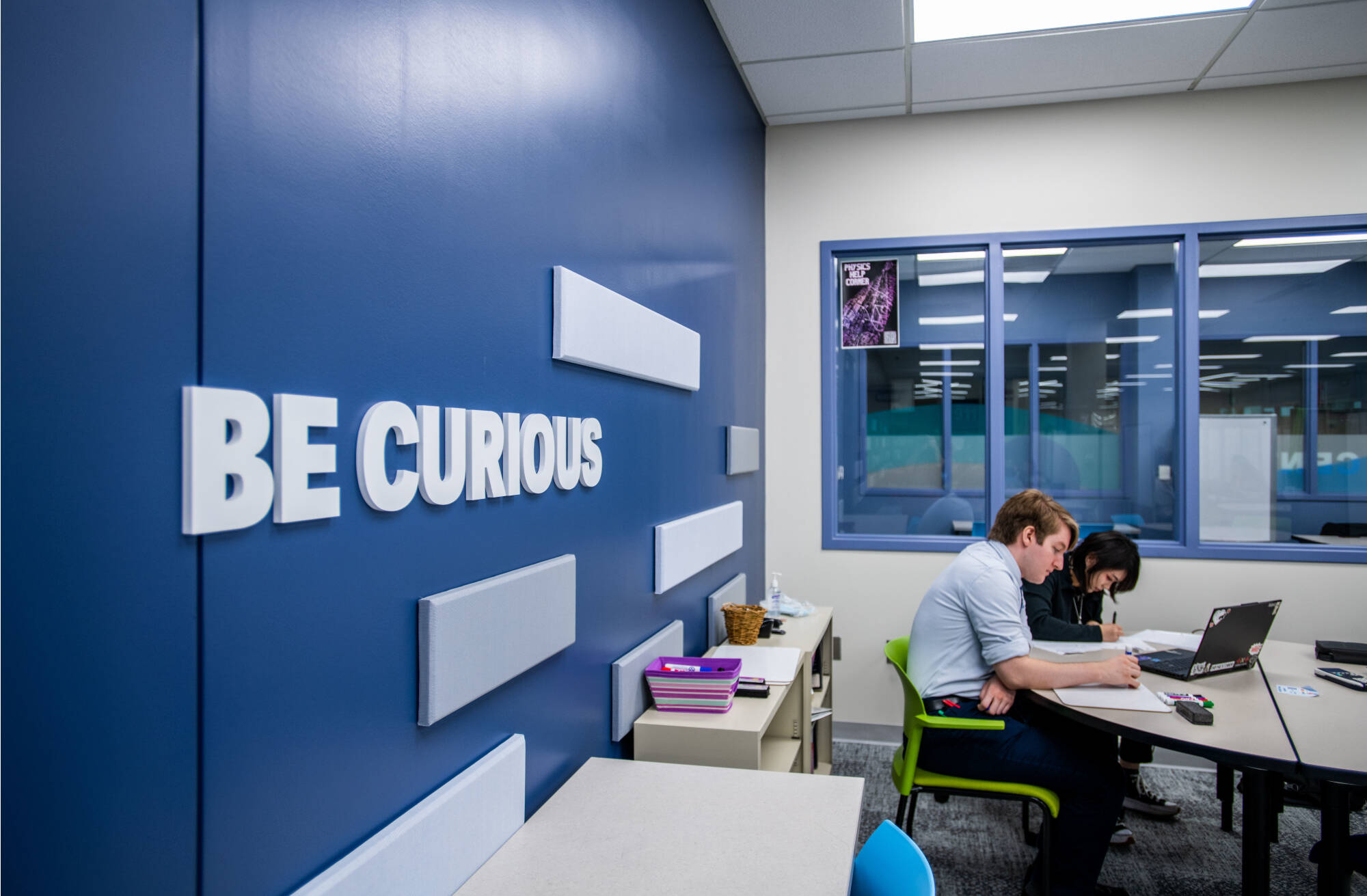 Two Students Studying in the Tutoring and Reading Center, with "BE CURIOUS" Written on a Wall Behind Them